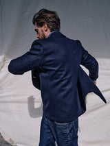A brown-haired person wearing a Blue de Gênes Peacoat - Dark Navy, featuring branded buffalo horn buttons, and jeans is photographed from behind against a light fabric backdrop.