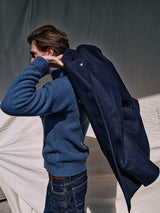 A person with dark hair is seen from the side putting on an Aimone Knit Cardigan in Sugar Blue by Blue de GĂȘnes over a coarse pearl knit sweater, paired with dark jeans, set against a backdrop of draped white fabric.