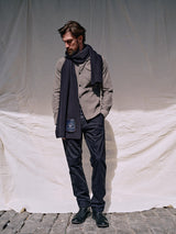 A person stands on cobblestone against a white fabric backdrop, wearing a beige jacket, Menzo Alvaro Chino in navy by Blue de Gênes with tapered legs, black shoes, and a dark scarf while looking down with a contemplative expression.
