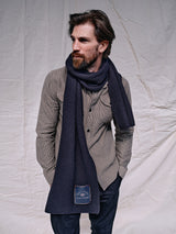 A bearded man in a Blue de Gênes Ginotto Oregon Shirt in pebble beige, paired with a dark scarf, stands against a light wrinkled backdrop. The classic fit brushed cotton shirt adds understated elegance as he gazes thoughtfully to the side.