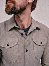 A bearded person wears the Blue de Gênes Ginotto Oregon Shirt in Pebble Beige, featuring a classic fit, yarn-dye stripes, brushed cotton fabric, and two chest pockets with black buttons. They look to the side against a plain light background.