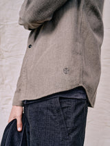 A person is wearing the Ginotto Oregon Shirt in Pebble Beige by Blue de Gênes, made of brushed cotton with a small emblem near the hem, paired with dark pants and holding a black item. The shirts texture stands out against a neutral fabric backdrop.