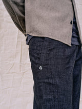A person sports a textured brown jacket over a black shirt and Menzo Alvaro Chino - Navy with a tapered leg, by Blue de Gênes, featuring a small diamond logo on the pocket. The background shows a beige, wrinkled surface for added comfort stretch ambiance.