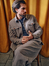 A man with dark hair and a beard sits on a chair in front of yellow curtains, exuding refined style in a taupe jacket over a blue button-up shirt and Blue de Gênes Paco Papago Chino in Stormy Grey, looking to the side with hands clasped.