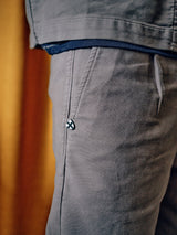 Close-up of a person in Blue de Gênes Paco Papago Chino in Stormy Grey, featuring a small logo near the pocket. The refined style is accentuated by the soft fabric and complemented by a dark blue shirt at the waistband. The background is a warm blur.