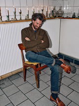 A bearded person with a mustache sits on a wooden chair in a tiled room, wearing the Aimone Knit Cardigan by Blue de GĂȘnes, jeans, and brown loafers. Their arms are crossed and theyâre smiling. Behind them, lit candles flicker gently on a shelf.