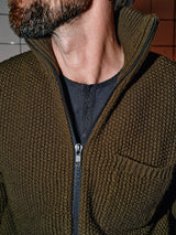 A close-up features a person in an Aimone Knit Cardigan - Army by Blue de GĂȘnes with its zipper partially open, exposing a black shirt underneath. The emphasis is on the knit texture, capturing some of the persons face and neck.