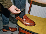 A person in leather loafers sits on a yellow chair, adjusting the rolled cuff of their Blue de Gênes Vinci Haworth Jeans in Denim Blue, featuring Comfort Stretch Denim. They don black socks and a knitted brown sweater, effortlessly stylish.