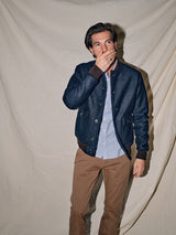 In front of a beige fabric backdrop, a man exudes a vintage look with his dark blue jacket, light blue shirt, and Blue de Gênes Menzo Dobby Chino - Brownie pants. His right hand covers his mouth as he gazes to the side, soft shadows from the lighting add depth to the scene.