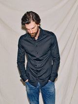 A man with dark hair and a beard wears the Blue de Gênes Luccio Dry Shirt in Jet Black and blue jeans. He stands against a light fabric backdrop, looking down with his hands in his pockets.