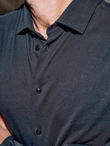 Close-up of a persons chest in the Blue de Gênes Luccio Dry Shirt - Jet Black, showcasing its garment-dyed textured pattern. The cotton pique jersey is slightly wrinkled, buttoned to the collar, with subtle logo embroidery. The image captures from neck down.