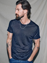 A bearded man with tattoos and a necklace, dressed in a Blue de Gênes Totti T-Shirt - Dark Navy and jeans, gazes to his right. The backdrop of draped light-colored fabric adds natural texture to the setting.