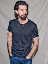 A bearded man with short dark hair stands against a plain backdrop, wearing the Totti T-Shirt in Dark Navy by Blue de Gênes, paired with jeans and a chain necklace. With hands in pockets and tattoos on his arms, he gazes right, showcasing the subtle depth of the 100% linen jersey.