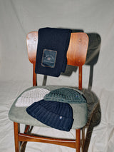A wooden chair with a navy scarf draped over the backrest, featuring three wool blend Oscar Melange beanies by Blue de Gênes in grey, navy, and green on the seat, showcasing 3-gauge knit details against a textured light backdrop.