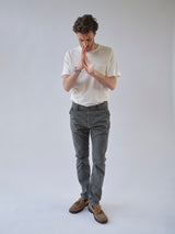 A man in a white t-shirt, BGpaulo Pavia 1 V2 Chino - Charcoal by Blue de Gênes, and brown shoes stands against a plain light background, looking down with his hands pressed together in front of his face as if deep in thought.