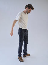 A man stands against a plain light background, wearing a white t-shirt, Blue de Gênes BGpaulo Pavia 1 V2 Chino in Midnight Blue, and light brown moccasin-style shoes. He looks down with a relaxed posture.