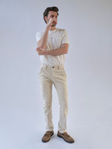 A man stands against a plain background, wearing a white stretch cotton t-shirt and Blue de Gênes BGpaulo Pavia 1 V2 Chino pants in ecru, with brown shoes. He crosses one arm and touches his face, gazing thoughtfully into the distance.
