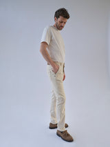 A man stands in a light studio wearing a white T-shirt, brown shoes, and Blue de Gênes BGpaulo Pavia 1 V2 Chino in Ecru, with his hands in his pockets and head tilted downward.