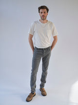 A man with short brown hair stands against a plain background, wearing a white t-shirt, Blue de Gênes BGrepi Giulio Light V2 Jeans in grey denim, brown loafers, and a silver necklace, hands in pockets and a neutral expression.