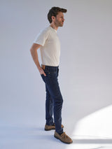 A man in profile wears a white t-shirt, brown shoes, and Blue de Gênes BGrepi Dai V Dark V2 Jeans in dark blue denim, with one hand in his pocket against a plain light background.