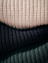 Close-up of the Blue de Gênes Yomaha Halfzip Knit in Navy, featuring a chunky ribbed texture, soft thick yarn, and a cozy wool blend. Sweater is stacked with others in beige and dark green.