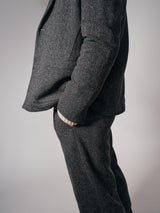 A person, shown from shoulders down against a plain background, wears refined tailoring—a grey herringbone suit with Blue de Gênes Kyoto Pant in Black Pattern—and has their hand in their pocket.
