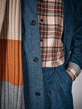 A person wearing a blue denim jacket and pants, the Blue de Gênes Robertino Brush Shirt in Stone, and a scarf with beige, orange, and gray stripes, with one hand in their pocket.