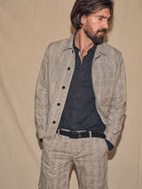 A bearded man in a gray checkered outfit, wearing a Blue de Gênes black Intero Belt over a dark shirt, stands against a plain beige background, looking right with his hands in his pockets.