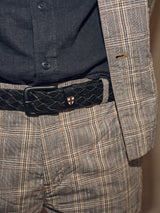 A close-up reveals a person in a plaid suit and dark shirt. The Intero Belt in black by Blue de Gênes, featuring a small shield emblem, accents the waist. Hand-sewn stitches enhance the beige and brown-toned suit, adding a vintage touch to the ensemble.