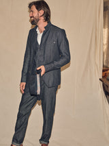 A man in the Casella Wasabi Blazer - Denim Blue by Blue de Gênes, paired with a white shirt and patterned tie, stands smiling and looking sideways. His medium-length hair and beard complement the cream-colored fabric backdrop. Buffalo horn buttons adorn the dark denim suit jacket.