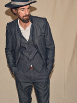 A bearded man wears a Casella Wasabi Blazer in Denim Blue by Blue de Gênes, paired with a white shirt and patterned hat. He stands against a beige backdrop, hands in pockets, looking slightly to the side.