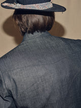 A person with shoulder-length brown hair is viewed from behind, wearing the Blue de Gênes Casella Wasabi Blazer in Denim Blue and a black hat featuring a paisley-patterned band. The blazers dry denim texture lends a rugged charm that beautifully enhances the ensemble.