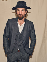 A man with a beard, donning a dark hat and textured suit, stands confidently against a neutral backdrop. His light shirt contrasts with his attire. Relaxed with hands in pockets, he looks at the camera—his outfit completed with the Pento Belt by Blue de Gênes.