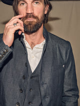 A bearded man in a Casella Wasabi Blazer - Denim Blue by Blue de Gênes over a white shirt thoughtfully touches his lips. The blazers buffalo horn buttons and high collar enhance its distinctive style.