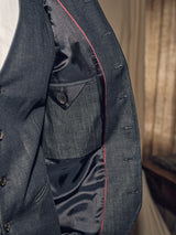 Close-up of a person wearing an open denim blue Casella Wasabi Blazer by Blue de Gênes, highlighting the inner lining with stitching details and a buttoned pocket. The jacket showcases red piping, buffalo horn buttons, and reveals a partially visible white shirt underneath.