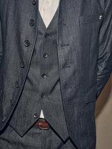 Dressed in a denim blue Casella Wasabi Blazer by Blue de Gênes over a crisp white shirt, they stand against a neutral background. The blazer features Buffalo horn buttons and a discreet logo on the pocket, with their belt subtly visible.