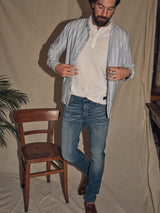 A man in blue jeans and a Sandro Caicos Shirt - Blue Stripe by Blue de Gênes, made from 6oz fine stripe fabric, stands near a wooden chair. Over a white top, the shirts Portuguese craftsmanship hints at Somelos origins. A potted plant adds charm against a beige fabric backdrop.