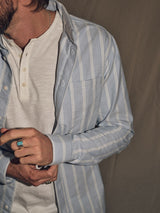 Wearing a Sandro Caicos Shirt in blue stripe by Blue de Gênes, layered over a white Henley and accessorized with a ring and chain necklace. The outfit, featuring fine stripe fabric made by Somelos in Portugal, is partly out of frame against a neutral backdrop.