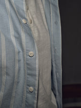 A person wears the Sandro Caicos Shirt - Blue Stripe by Blue de Gênes, crafted from Somelos in Portugal. The focus is on the buttons and fabric texture of the blue and white 6oz fine stripe, layered over a light gray t-shirt, against a dark, blurred background emphasizing craftsmanship.