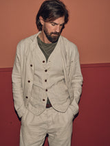 A man with long hair and a beard stands against a two-tone red-orange wall, sporting a relaxed look in the Blue de Gênes Gabbiano Winnot Blazer - Natural. He exudes effortless style with a green shirt under his beige double-breasted suit, one hand in his pocket and eyes cast downward.