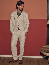 A bearded man leans against a red and orange wall, wearing the Blue de Gênes Gabbiano Winnot Blazer in natural herringbone over a green shirt, with brown shoes. With one hand in his pocket, he gazes sideways on a wooden floor beside a table edge.