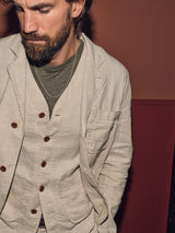 A person wearing a Blue de Gênes Gabbiano Winnot Blazer - Natural over a green shirt and vest with brown buttons stands against a two-toned wall, one hand in their pocket, looking down to the left.