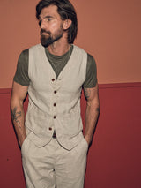 A tattooed and bearded man stands against a two-tone wall, exuding effortless style. Sporting Blue de Gênes Totti T-Shirt in Army under a beige vest with beige pants, he keeps his hands casually in his pockets while gazing to the side.