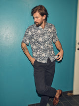 A bearded man poses against a teal wall in a floral shirt, Franco Genes Chino - Navy by Blue de Gênes, and brown shoes. With one hand in his pocket, his body slightly turned, one leg is crossed in front of the other.