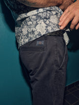 A person wearing a floral shirt and Franco Genes Chino - Navy by Blue de Gênes stands against a teal wall. Their hand rests on their hip, revealing part of a patterned belt.