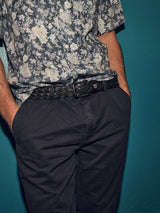Against a teal backdrop, someone is styled in a navy blue and white floral shirt with Franco Genes Chino - Navy by Blue de Gênes. Their hands rest casually in pockets, complemented by a black woven belt at the waist.