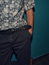 A person wearing a floral-patterned shirt with rolled-up sleeves and Blue de Gênes Franco Genes Chino in Navy stands against a turquoise wall. Their right hand is in their pocket, revealing a black woven belt.