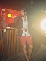 A man with a beard stands at a wooden bar holding a drink, wearing a striped shirt, Teo Milano Shorts - Havana Red by Blue de Gênes, and a dark blazer. The warm, hazy lighting creates a relaxed atmosphere.