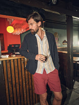 A man with a beard stands by a bar, drink in hand. He wears a dark blazer over a striped shirt and vibrant Teo Milano Shorts in Havana Red by Blue de Gênes, made from a comfy cotton/linen blend. The wooden counter and hanging light create an ideal setting for relaxation.