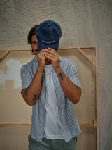 A tattooed man hides his face under a blue cap while wearing a striped short-sleeve shirt over the Blue de Gênes BdG S/S Grandad t-shirt in Snow White, styled with cotton slub yarn, against a neutral backdrop.